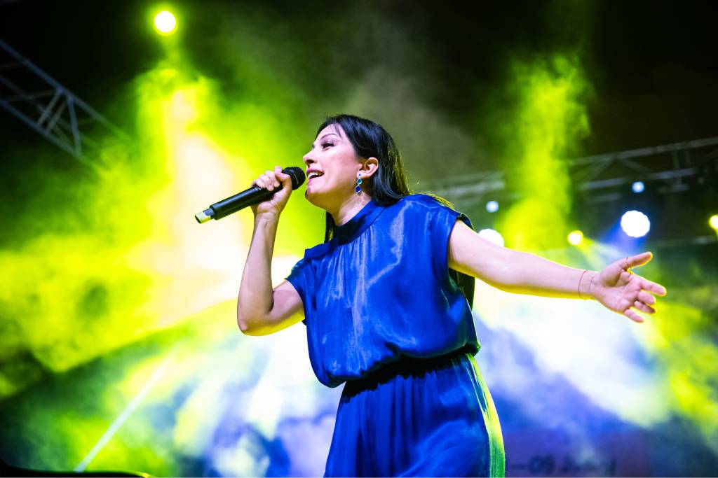 Mais Harb singing into a handheld microphone on stage in a blue dress under green stage lighting