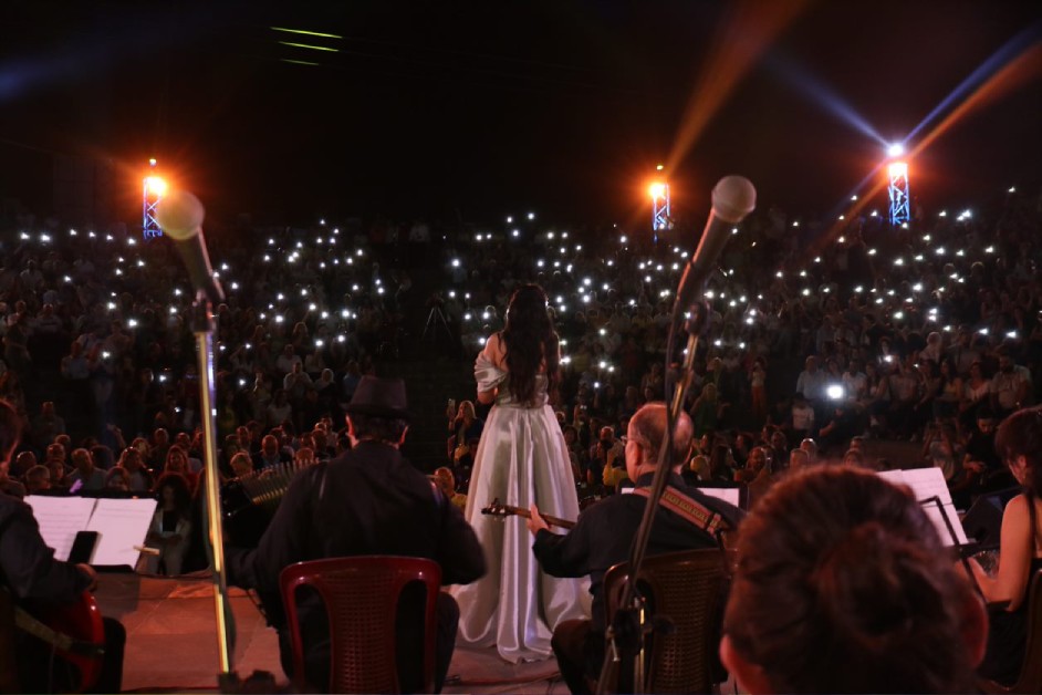 Mais Harb performing in a white gown before a large outdoor audience holding up phone lights, with her band on stage